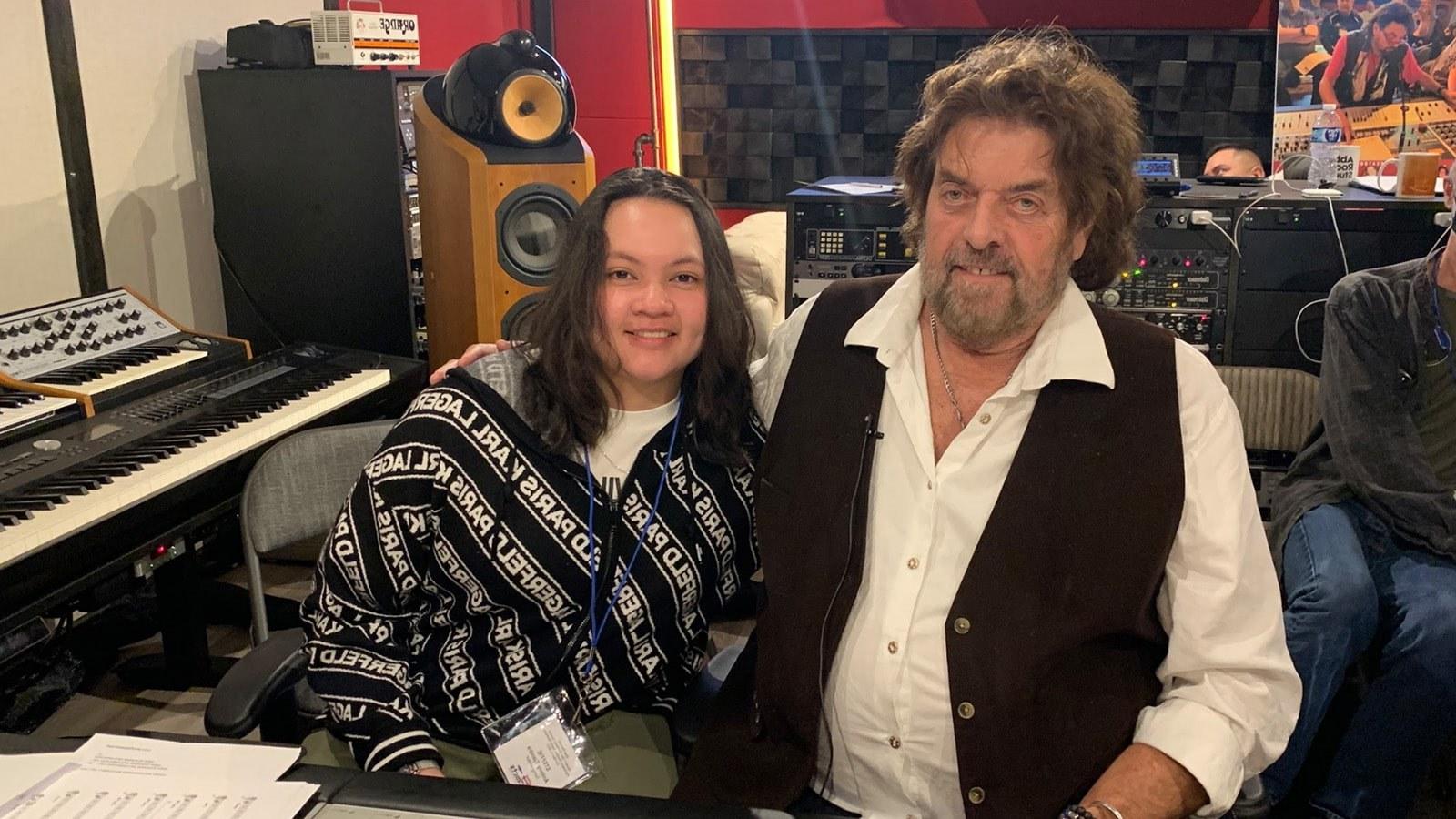 Alan Parsons sits with Steffie Tjandra in front of a mixing board in a recording studio. They are smiling at the camera.